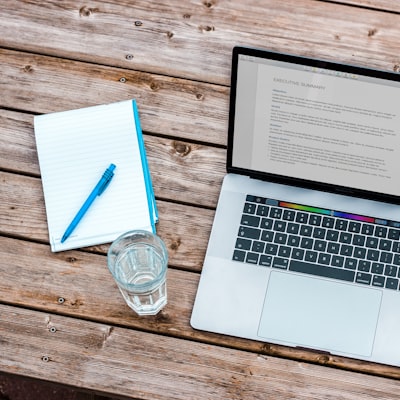 Laptop on a wooden table with an open document on screen, a blue pen on a notebook, and a glass of water nearby.